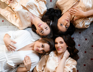 Above view of thoughtful and pretty bride with her friends, which wearing in white and beige silk peignoirs, lying on floor in room, sexually looking and posing at camera during wedding day