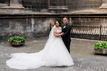 Front view of cute bride in wedding gown with long train and veil, standing near groom , holding his hand, looking and posing at camera during wedding walk in ancient city