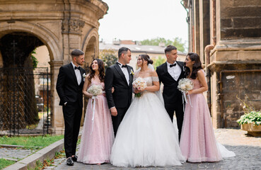 Front view of beautiful bride and groom in wedding apparel, smiling and looking to each other while standing between friends. Pretty bridesmaids with curly long hair, dressed in pink evening gowns