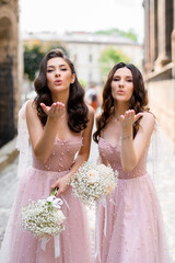 Naklejka premium Front view of two sexual girls with curly hairdo, wearing in long pink evening dresses, which adorned by pearls, holding bouquets with white roses and gypsophila, sending air kiss while posing