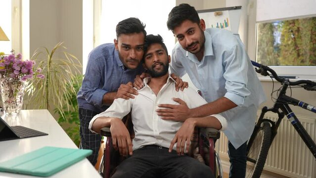 Smiling friends hugging man in wheelchair looking at camera sitting indoors in home office. Cheerful Middle Eastern mates supporting intelligent motivated friend posing