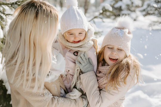 Portrait of woman playing with children during winter walk. Happy little girls, wearing warm jackets and scarfs, having fun with mother and playing with snow in forest