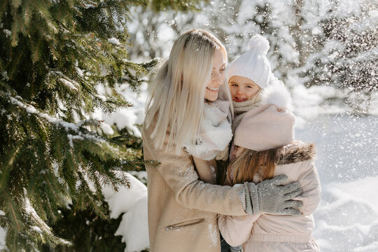 Portrait of mother spending time with children during winter holiday in snowy park. Smiling woman, dressed in warm outwear, embracing her daughters, while standing near tree during light snow falling