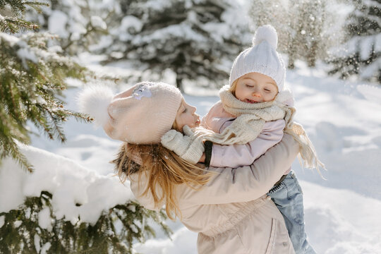 Portrait of loving little sisters embracing in a snowy forest. Little girl, spending winter holiday with sister, standing in background of piles of snow and trees in forest during sunny day