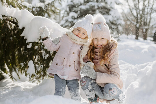 Portrait of cute little girls posing in snowy forest. Happy sisters, wearing warm clothes and scarves, holding with hands and standing in background of sunny snowy park