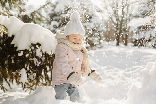 Portrait of cute child playing with snow in the forest. Funny little girl in warm jacket and scarf around neck, enjoying of play with snow, standing alone among snowy trees in park