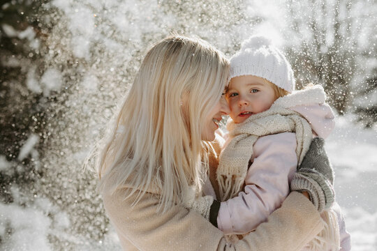 Portrait of mother holding cute little daughter, enjoying cold sunny weather in winter park. Attractive woman with blond hair, and braces on teeth, cuddling with little child
