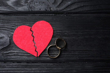 Divorce concept. Broken red paper heart and wedding rings on black wooden table, flat lay with space for text