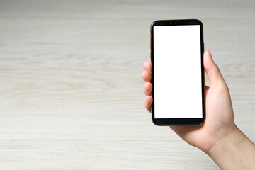 Woman holding smartphone with blank screen at white wooden table, top view. Mockup for design