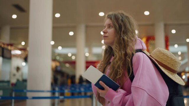 Pretty girl holding a ticket in abroad passport at international airport smiling and looking around. Young female tourist with a backpack and straw hat in the airport with a passport in her hands.