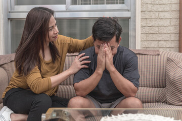 A depressed man breaks down cries while sitting on the sofa, covering his face while being comforted by his concerned wife. A supportive spouse being there in hard times.