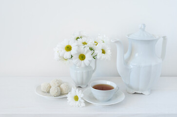 Obraz na płótnie Canvas Tea and flowers. A bouquet of white daisies or chamomile iflowers in a vase on the table, a cup of tea, sweets and a teapot on a white background