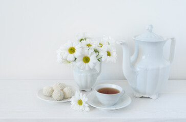 Tea and flowers. A bouquet of white daisies or chamomile iflowers in a vase on the table, a cup of tea, sweets and a teapot on a white background