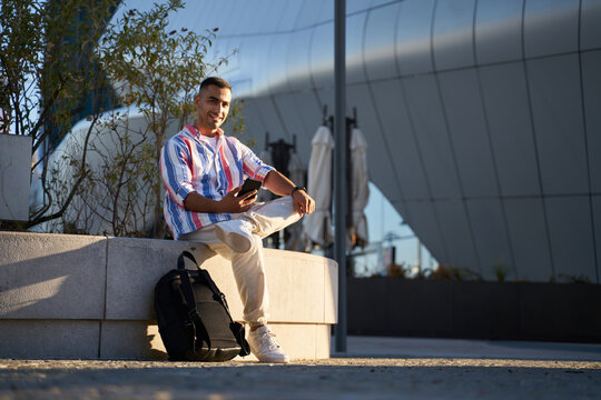 Young Iranian man holding smartphone and looking at the camera at the city. Handsome young guy in casual clothes relaxing outdoors at the city street 