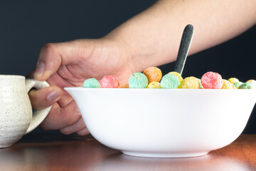 A man holding a small cup next to a deep plate filled with colorful sugary cereal on a wooden table