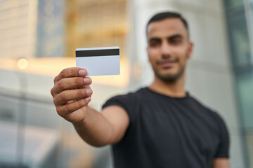 Handsome smiling middle eastern man holding credit card looking at camera, selective focus on card. Shopping concept