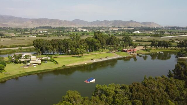 Drone rises above trees to reveal a large lake or lagoon in the middle of green agricultural fields. The lake is used for water sports. Recorded in 4k quality. Located in Ca&ntilde;ete in Southern Peru.