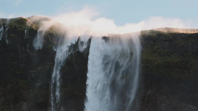 Mist from reverse waterfall in Iceland blows over top of majestic cliff.