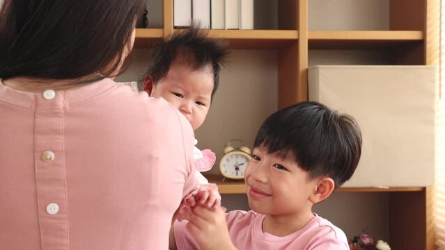 Asian boy excited with newborn girl wants to play with infant while mother hold in arm, sibling brother play together with love in bedroom having fun and cheerful, smiling boy looking to younger siste