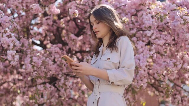 Casually dressed young woman taking selfie on smartphone while standing near sakura tree with lush spring flowering. Cute lady taking photo for social media. Modern gadgets for lifestyles