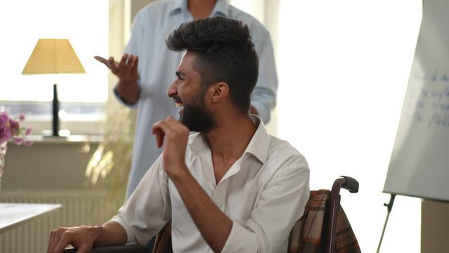 Joyful young man in wheelchair laughing out loud with unrecognizable friend gesturing standing at background. Cheerful positive Middle Eastern guy having fun with mate indoors