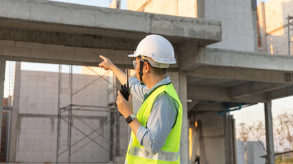 Skilled engineer inspects factory construction and uses a radio to chat with colleagues working on the field, Engineer working outdoors with blueprints and wearing protective safety gear.