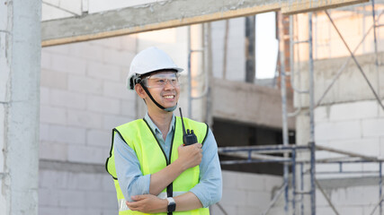 Skilled engineer inspects factory construction and uses a radio to chat with colleagues working on the field, Engineer working outdoors with blueprints and wearing protective safety gear.