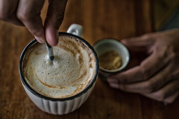 Hands mixing milk in hot coffee cup on wooden table in cafe.