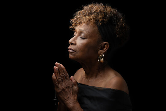 Beautiful elderly 70+ year old Christian African American black woman religiously and fervently praying to God with eyes closed and hands together holding a cross