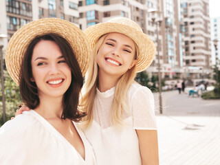 Two young beautiful smiling hipster female in trendy summer white dress clothes and straw hats. Sexy carefree women walking in street. Positive models having fun, hugging and laughing