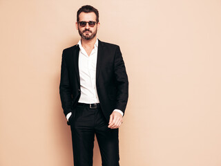 Fototapeta premium Portrait of handsome confident stylish hipster lambersexual model. Sexy modern man dressed in black elegant suit. Fashion male posing in studio, isolated on beige. In sunglasses