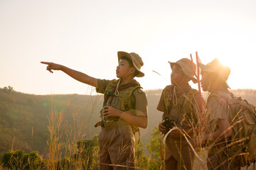 Asian boy scout schoolgirl hiking with backpack standing in the grass Three Boy Scouts Look into the distance to explore the beautiful forest in the evening as the sun sets. - obrazy, fototapety, plakaty