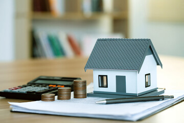 The house model and coins are stacked on the contract document, with a calculator beside them. Home...