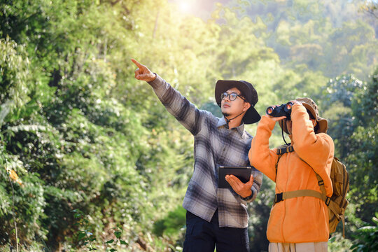 Asian woman backpacker nature explorer wearing hat and backpack Talk to male tourists holding binoculars during trekking explore while walking in nature forest with holiday joy