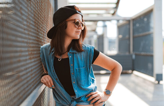 young stylish hipster girl in a daring image in a cap and sunglasses poses in the city in the rays of the sunset on an urban background.