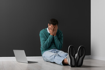 Upset man sitting on floor near laptop against black wall