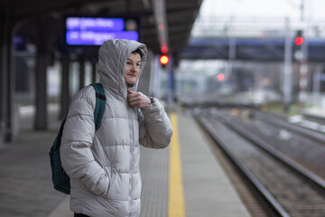 Fototapeta premium Thoughtful young man in casual gray jacket and hat with backpack standing and waiting for train at railway station.