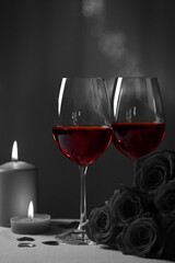 Glasses of wine, burning candles and rose flowers for romantic dinner on grey table against blurred lights. Black and white effect with red accent