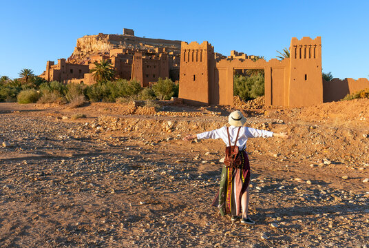 Beautiful young woman happy to travel in Morocco. Ait-Ben-Haddou kasbah on background. Wearing in white and hat