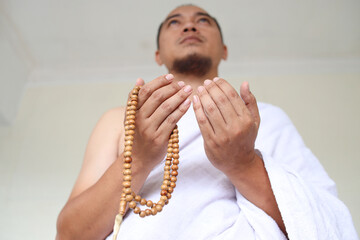 Low angle shot of Asian muslim man wearing white ihram clothes and praying while holding prayer beads - obrazy, fototapety, plakaty