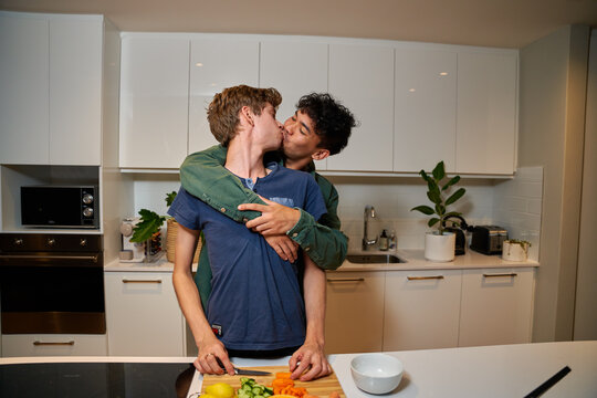Young gay couple in casual clothing kissing while preparing dinner in kitchen at home