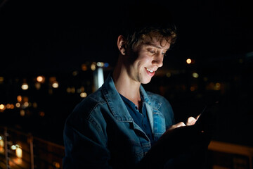 Young caucasian man in casual clothing smiling while using mobile phone on balcony at night