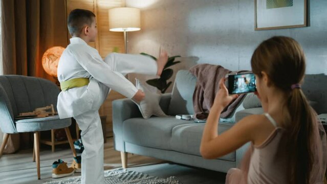 A little girl films a taekwondo boy doing some kicks in a room