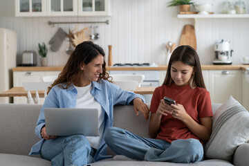Young smiling mother talking to teen daughter child about being online, modern family using digital devices while resting on sofa at home. Mom chatting speaking with kid about internet safety