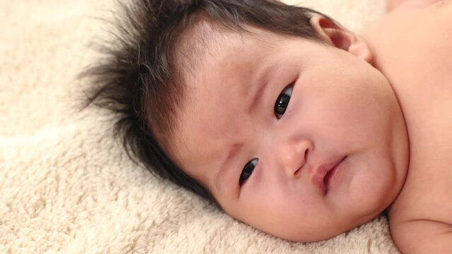 Close-up face asian newborn baby wakes up looking at camera and look around with doubt on bed in morning at home, adorable infant early days who wakes up and open his eyes. Concept of caring children.
