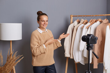 Delighted cute optimistic woman stylist or designer wearing beige sweater, presenting her new collection, shooting video for vlog, standing against gray wall with clothes hang in wardrobe on rack.