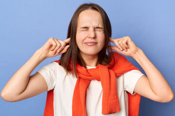 Frustrated annoyed woman in T-shirt and sweater tied over shoulders, covering ears with hands,...