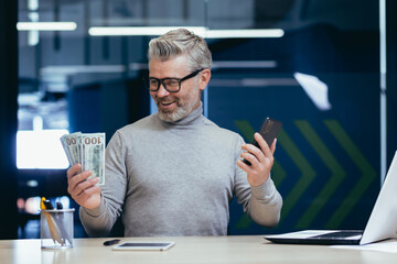 Mature gray haired businessman inside office happy with success and earned money, boss holding dollar cash and mobile phone smiling, senior investor successfully invested and earned.