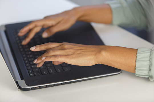 Hands of young adult black woman working on laptop behind white desk in the office. Overhead photo of BIPOC female person typing text on computer