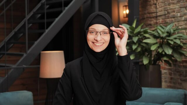 Portrait of beautiful pretty Arab muslim woman in black traditional hijab looking straight to camera and smiling in modern loft office. Close up of female pretty face with smile. Arabian headscarf.